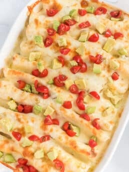 Rotisserie chicken enchiladas in a baking dish topped with avocado and tomatoes.