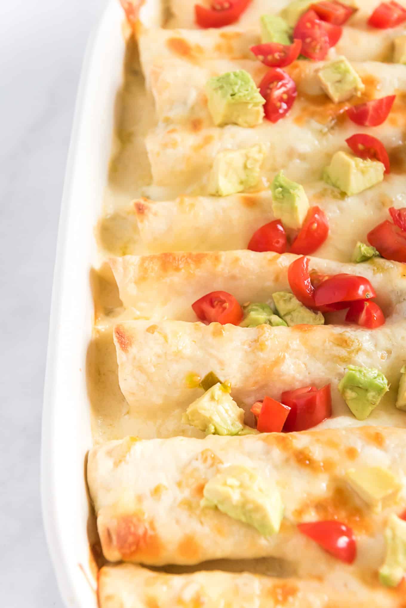 Rotisserie chicken enchiladas in a white casserole dish with tomato and avocado on the top.