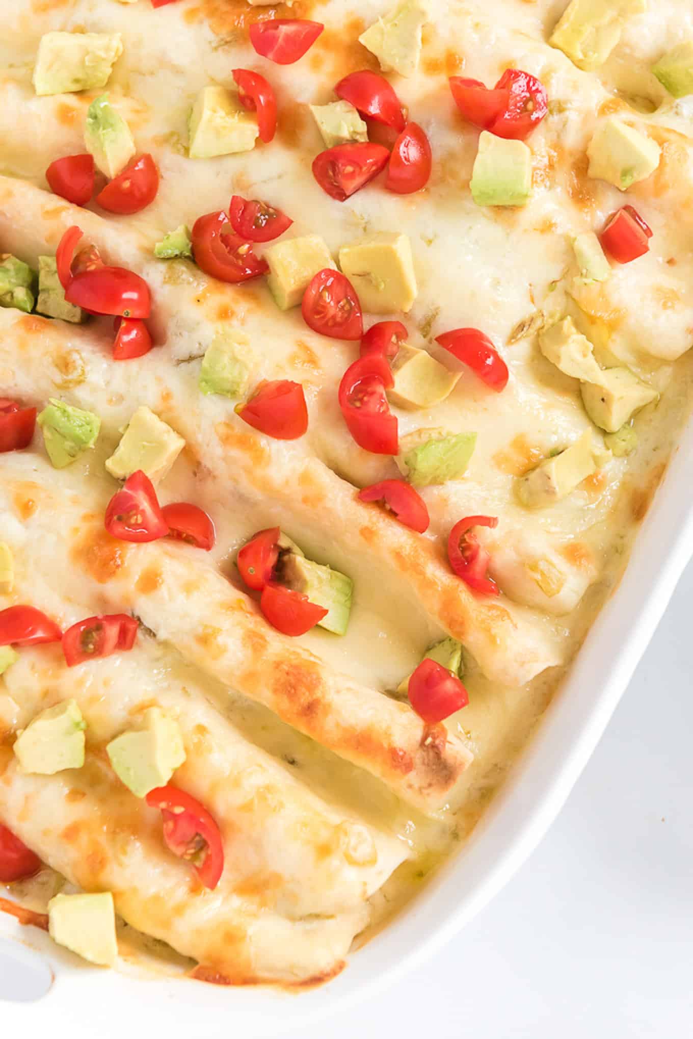 Rotisserie chicken enchiladas topped with melted cheese, tomatoes, and avocado in a baking dish.