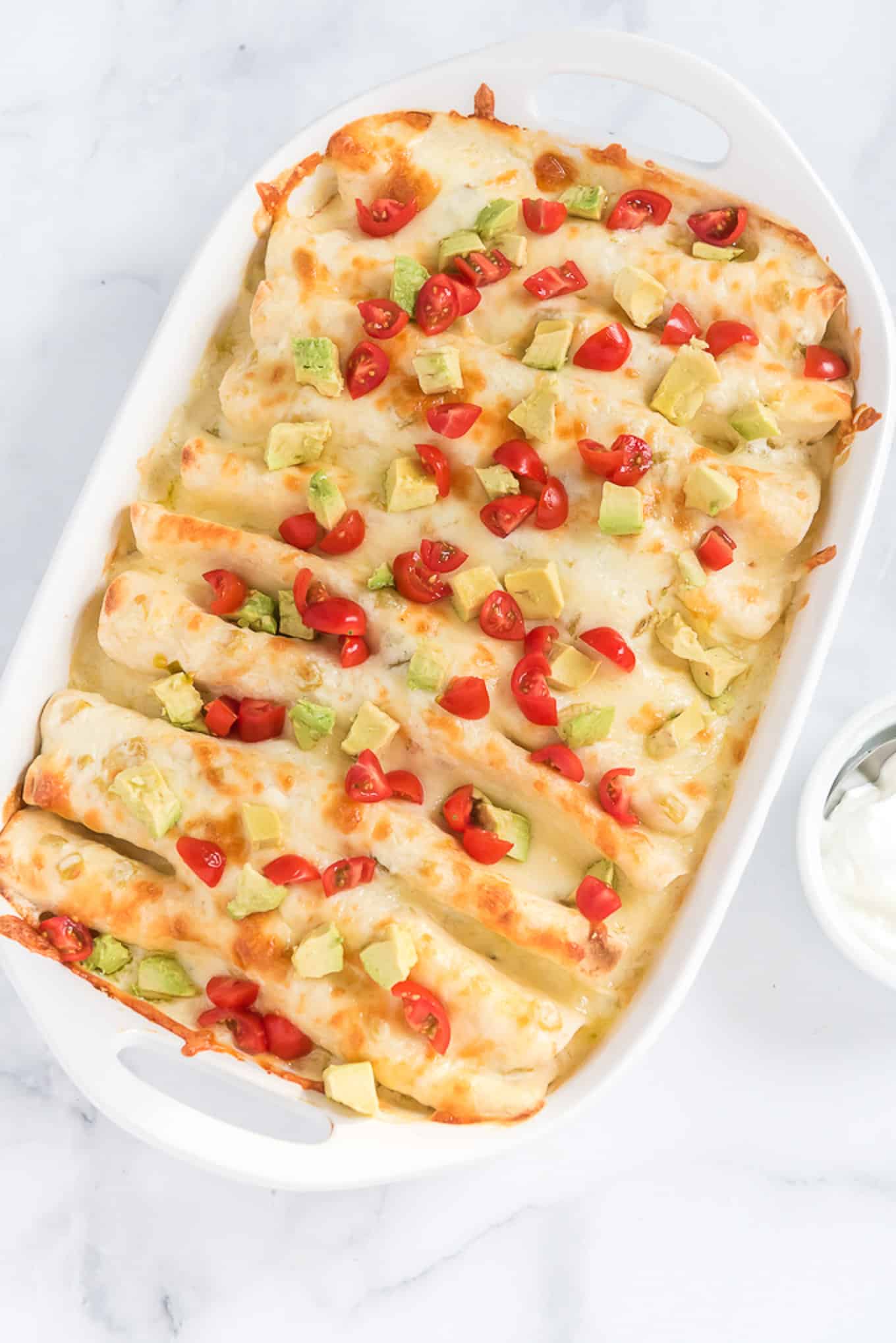 Creamy chicken enchiladas in a baking dish topped with tomatoes and diced avocado.