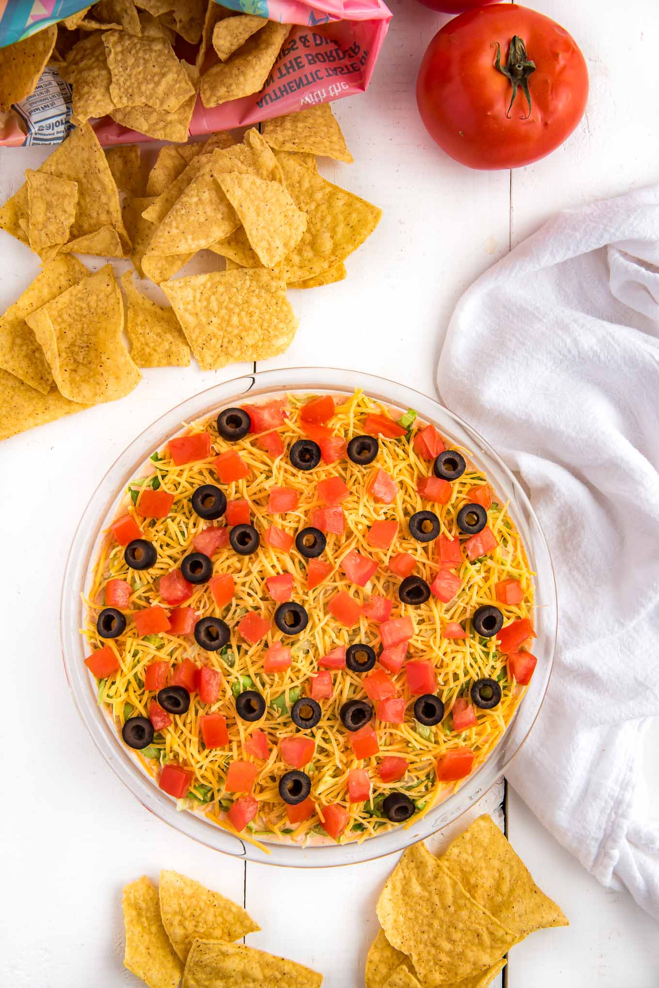 A serving bowl of taco dip on the table with a bag of chips spilling out next to it and a tomato.