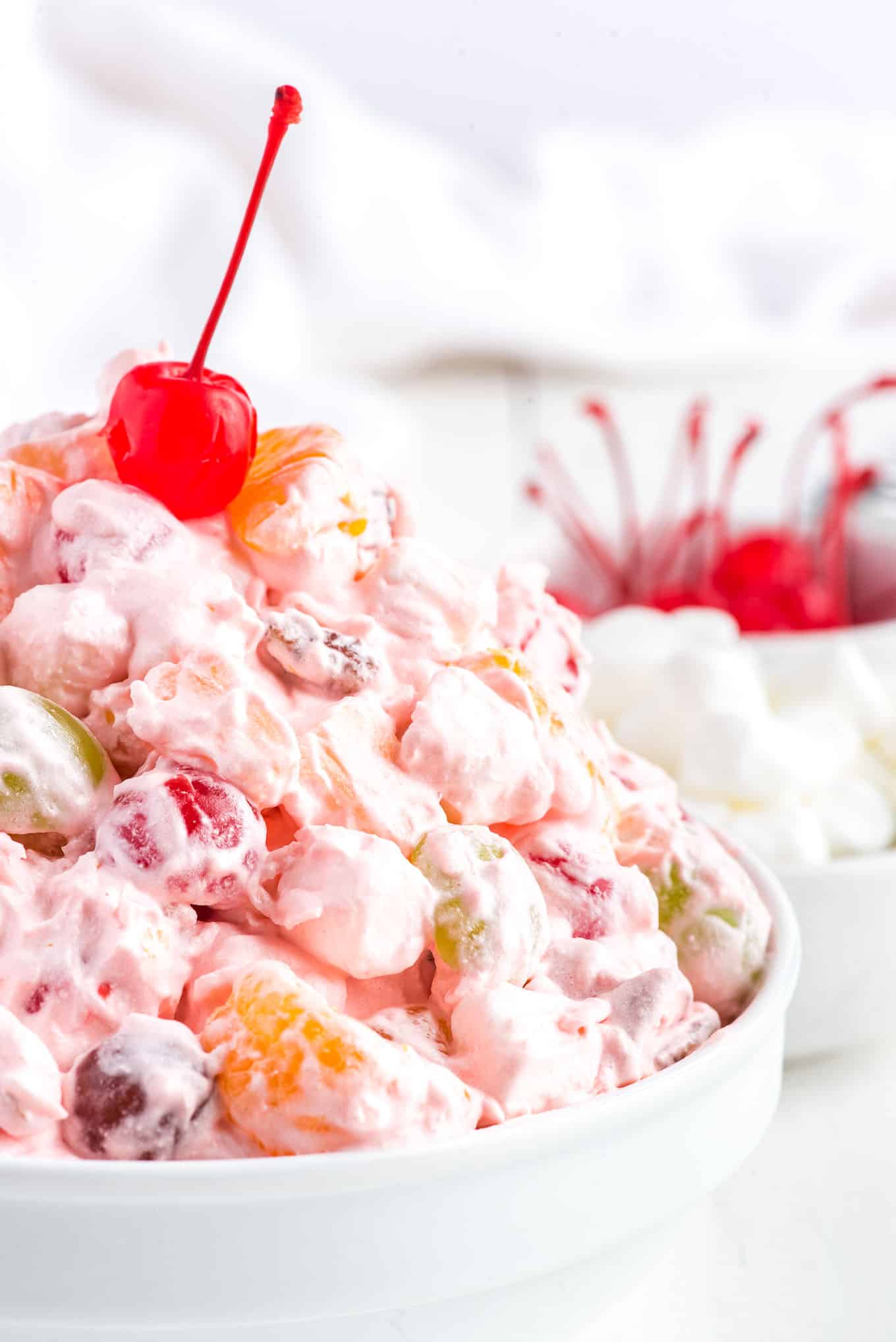 A bowl of fruit salad with Cool Whip on the table in front of a bowl of marshmallows.