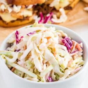 A bowl of coleslaw for pulled pork on the table in front of a platter of sliders.