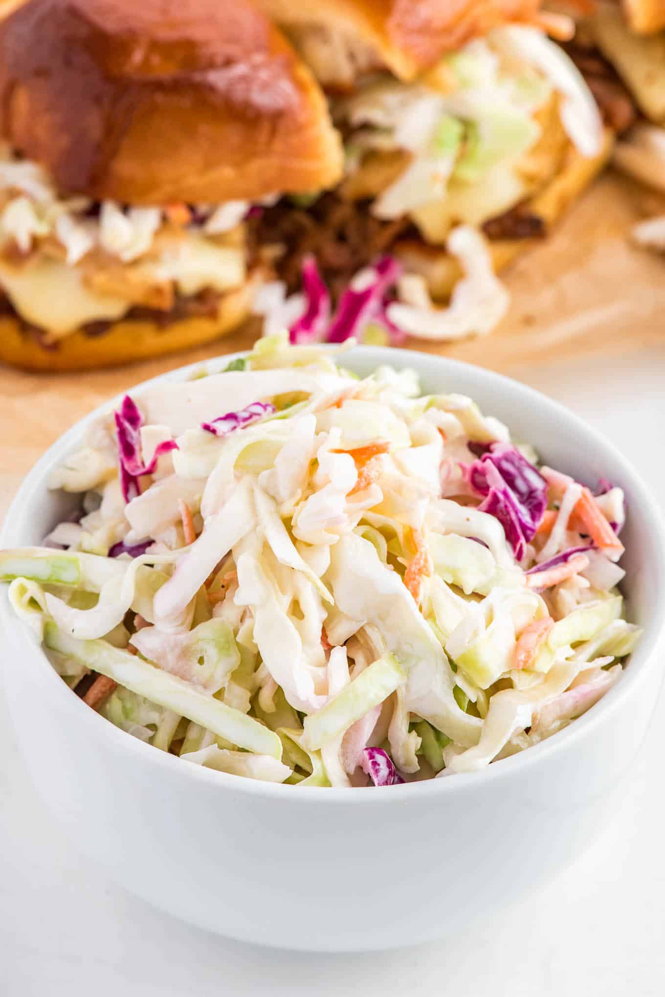 Coleslaw for pulled pork in a bowl in front of pork sliders in the background.