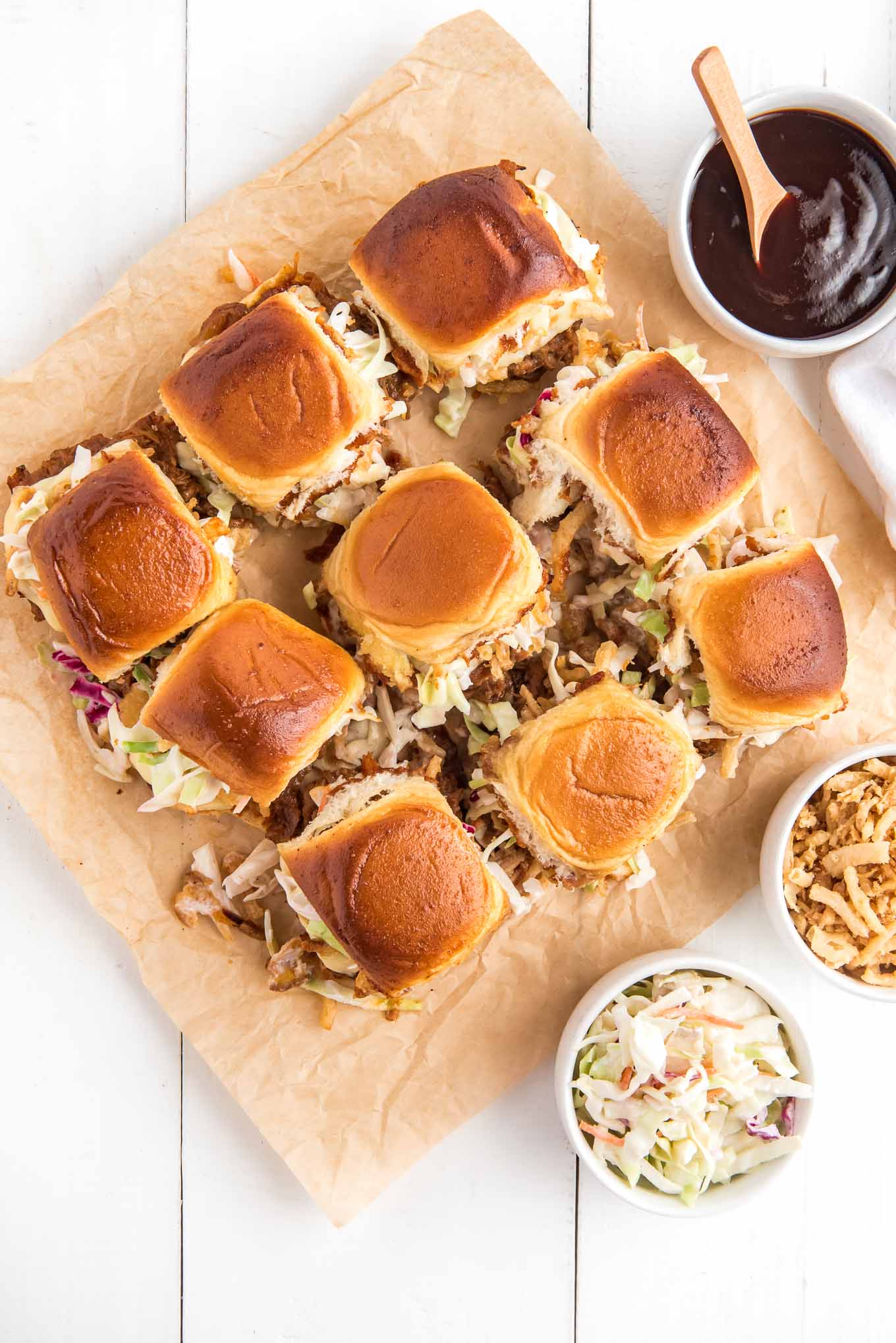 Pulled pork sliders on parchment paper with bowls of cole slaw, crispy onions, and bbq sauce around it.