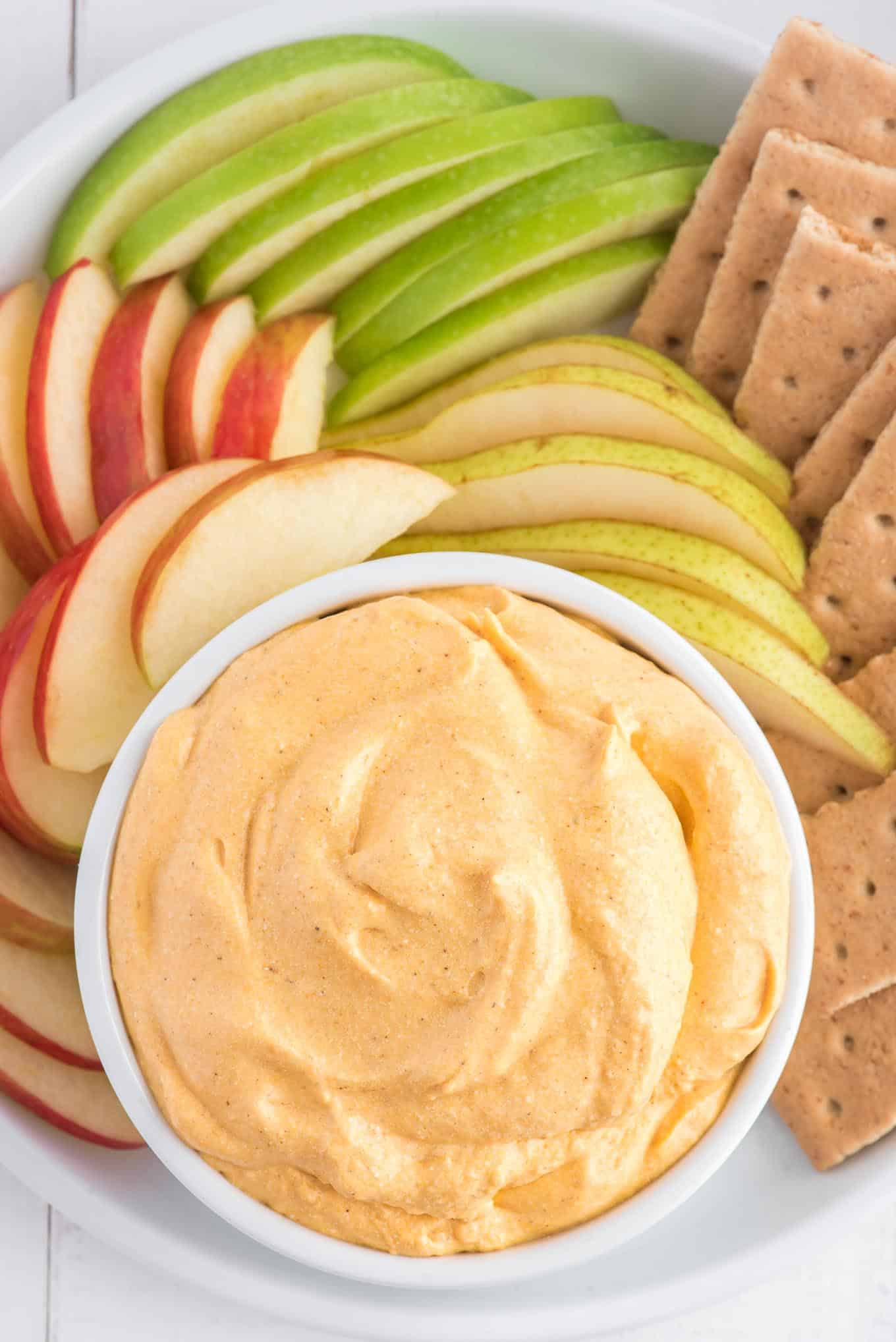 A bowl of pumpkin dip on a platter surrounded by apple slices, pear slices, and graham crackers.