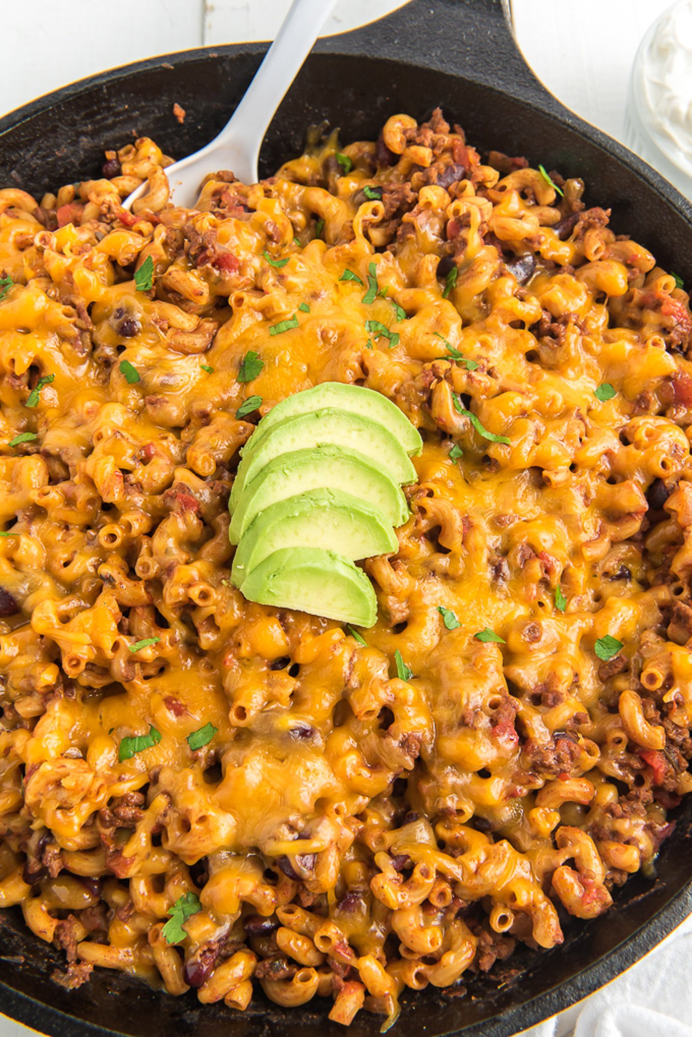 Chili mac in a skillet with a spoon scooping in and sliced avocados on top.