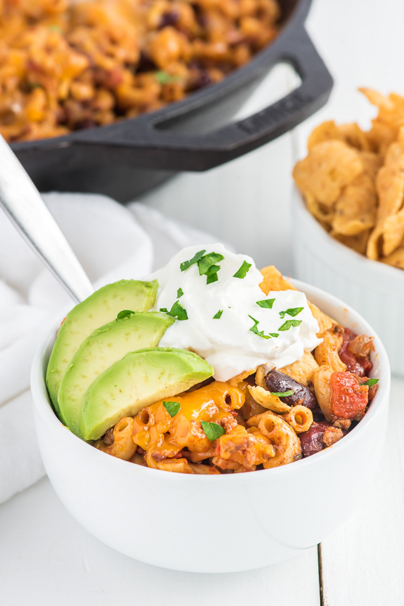 A bowl of chili mac on the table topped with sour cream and avocado with the skillet in the background.