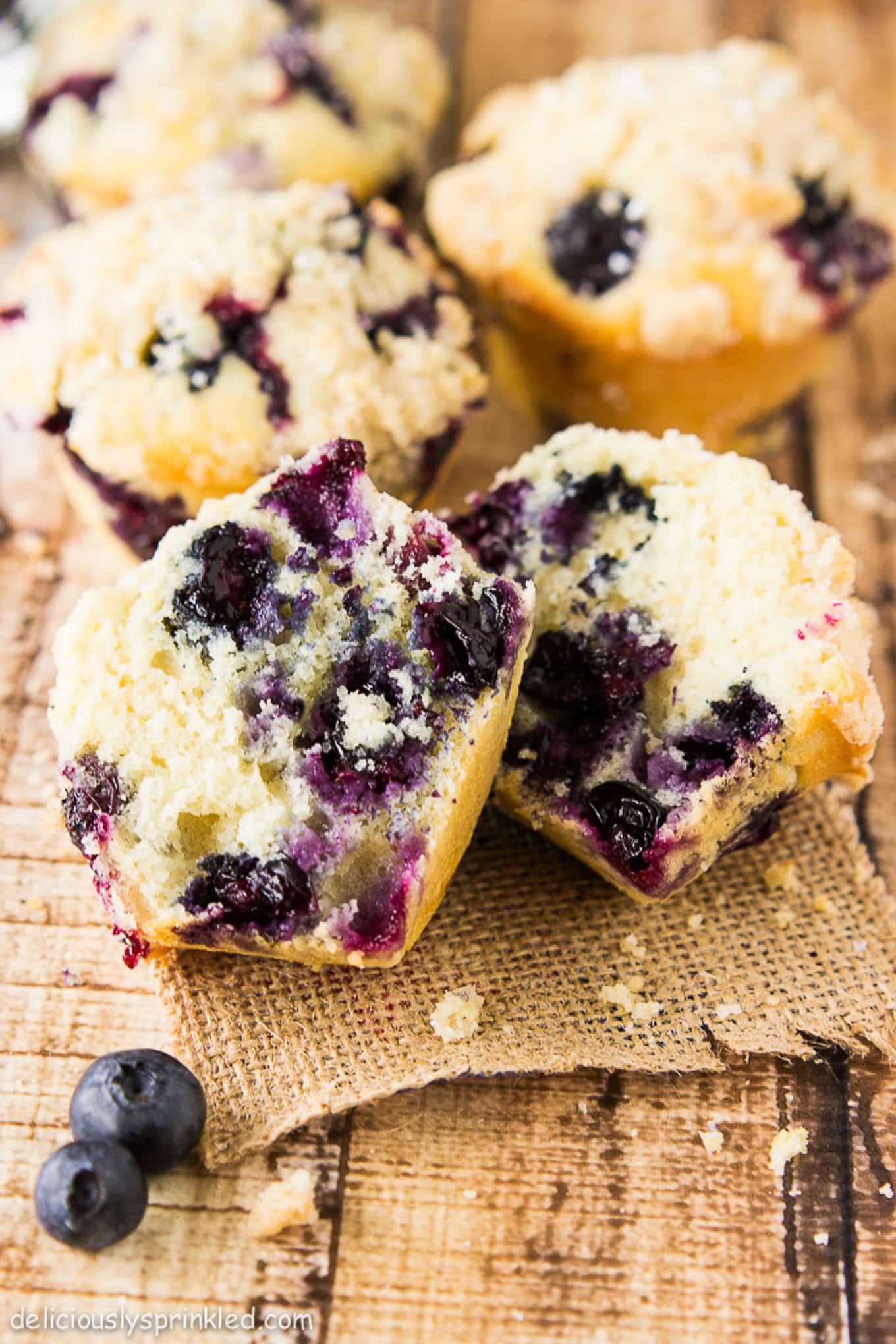 Homemade blueberry muffins on a burlap lined table with one cut in half to show the inside.