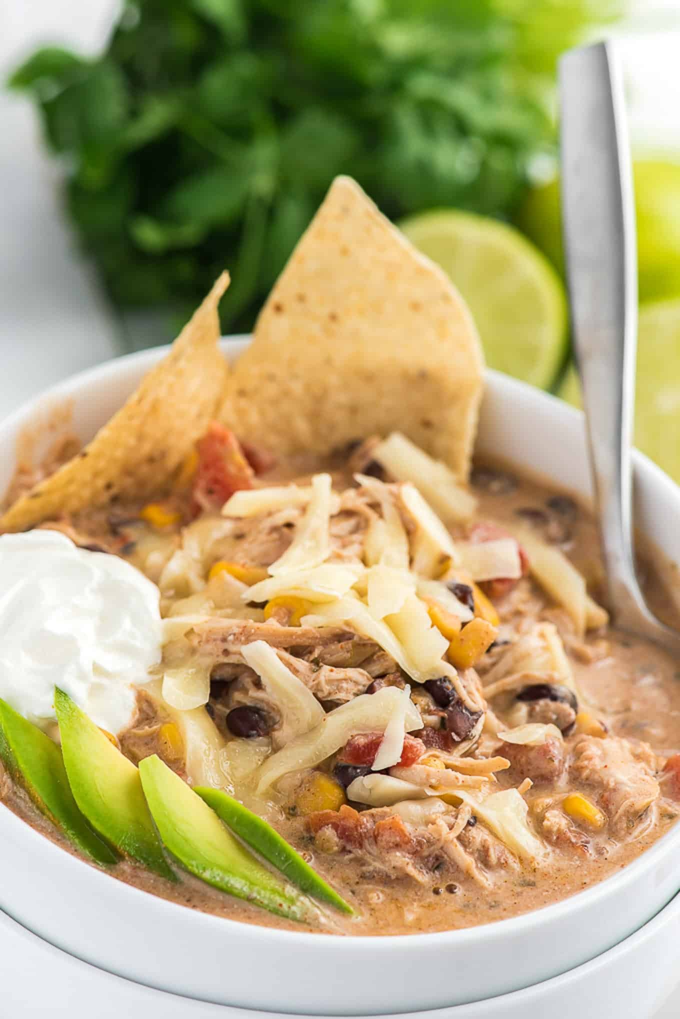 A bowl of white chicken chili on the table topped with cheese, sour cream, and tortilla chips.