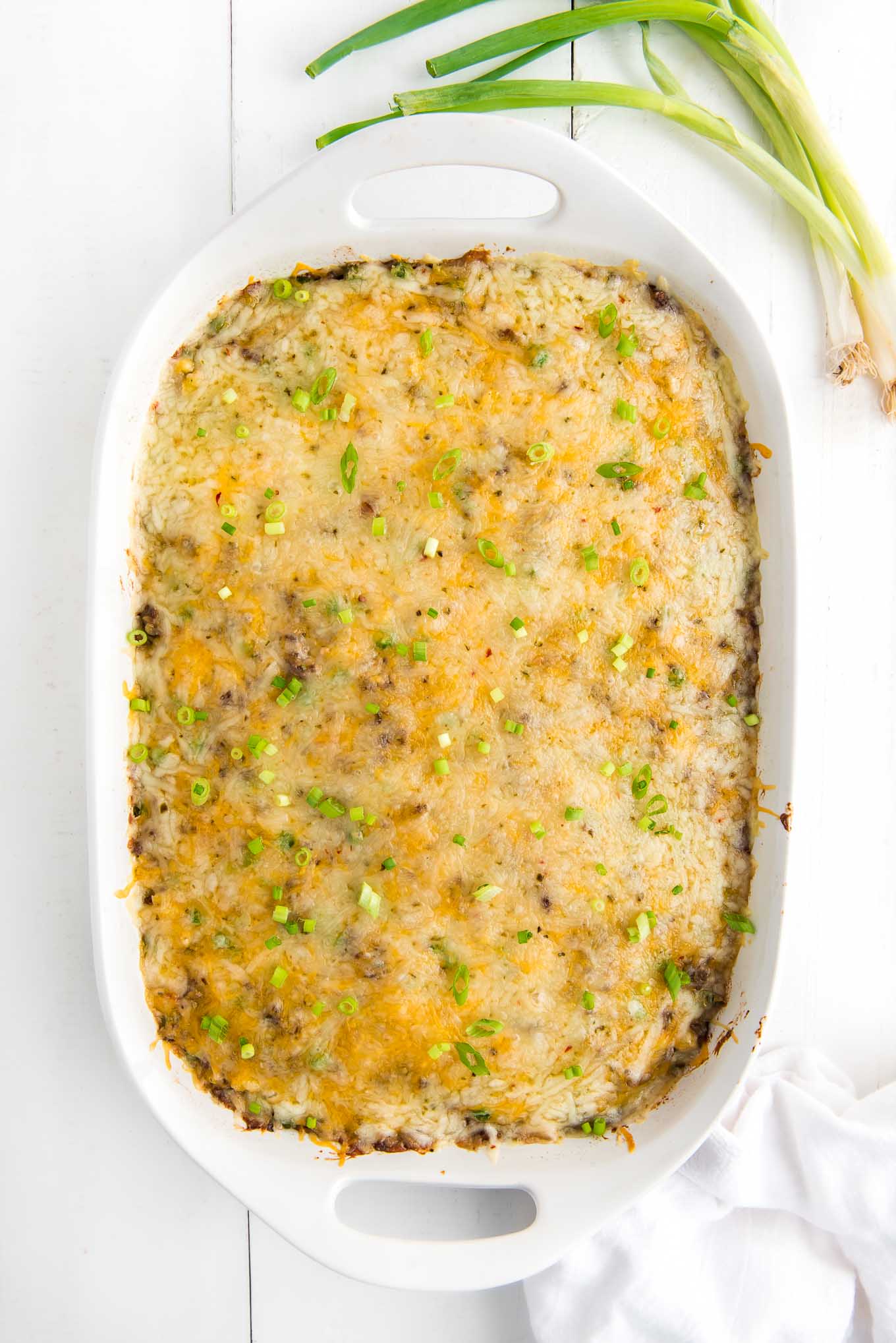 A baked hash brown casserole with hamburger topped with cheese and garnished with green onions on the table in a white rectangle dish.