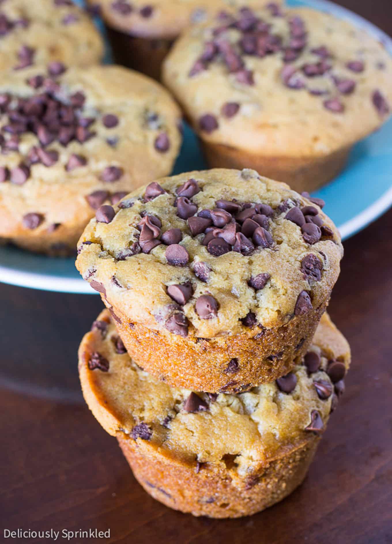 Two chocolate chip muffins stacked on the table with a plate of muffins in the background.