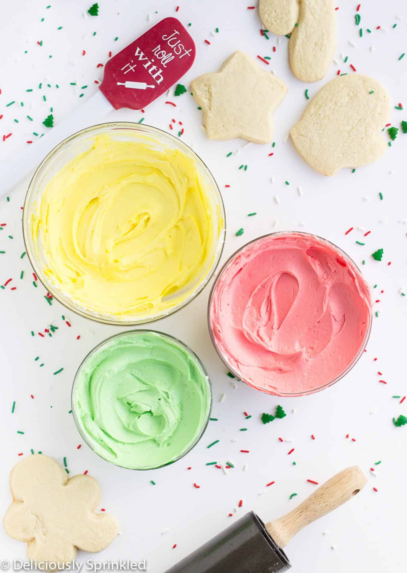 Bowls of colored cookie frosting in three different bowls on a white table with sprinkles and a few cookies scattered around.