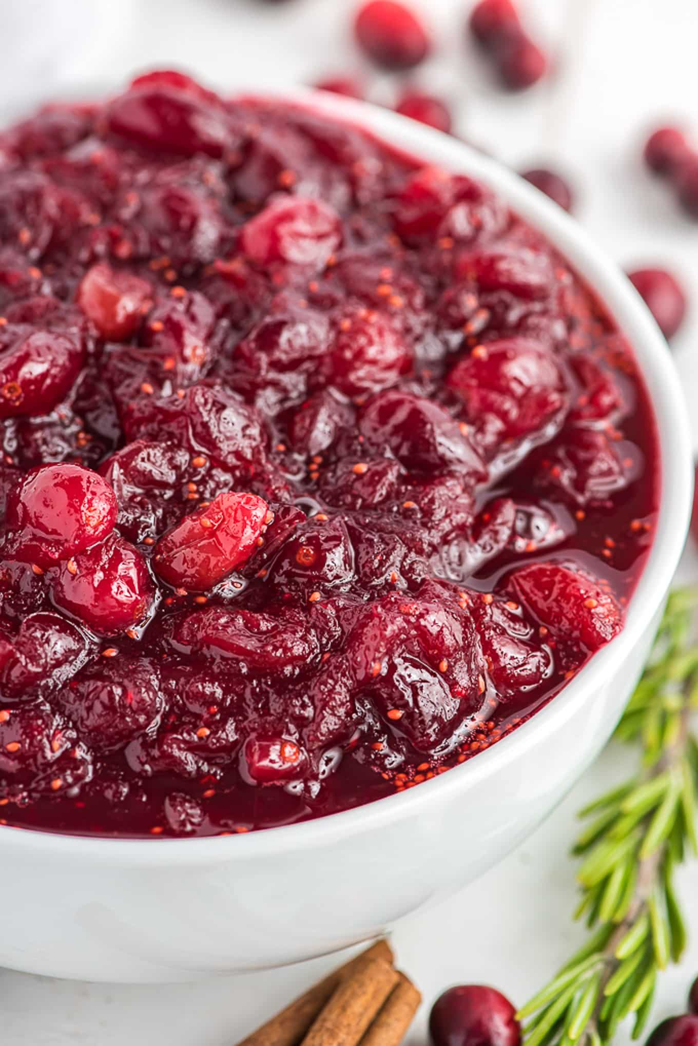 A closeup of a bowl of cranberry sauce ready to serve with sprigs of rosemary, cinnamon sticks and fresh cranberries on the table under the bowl.