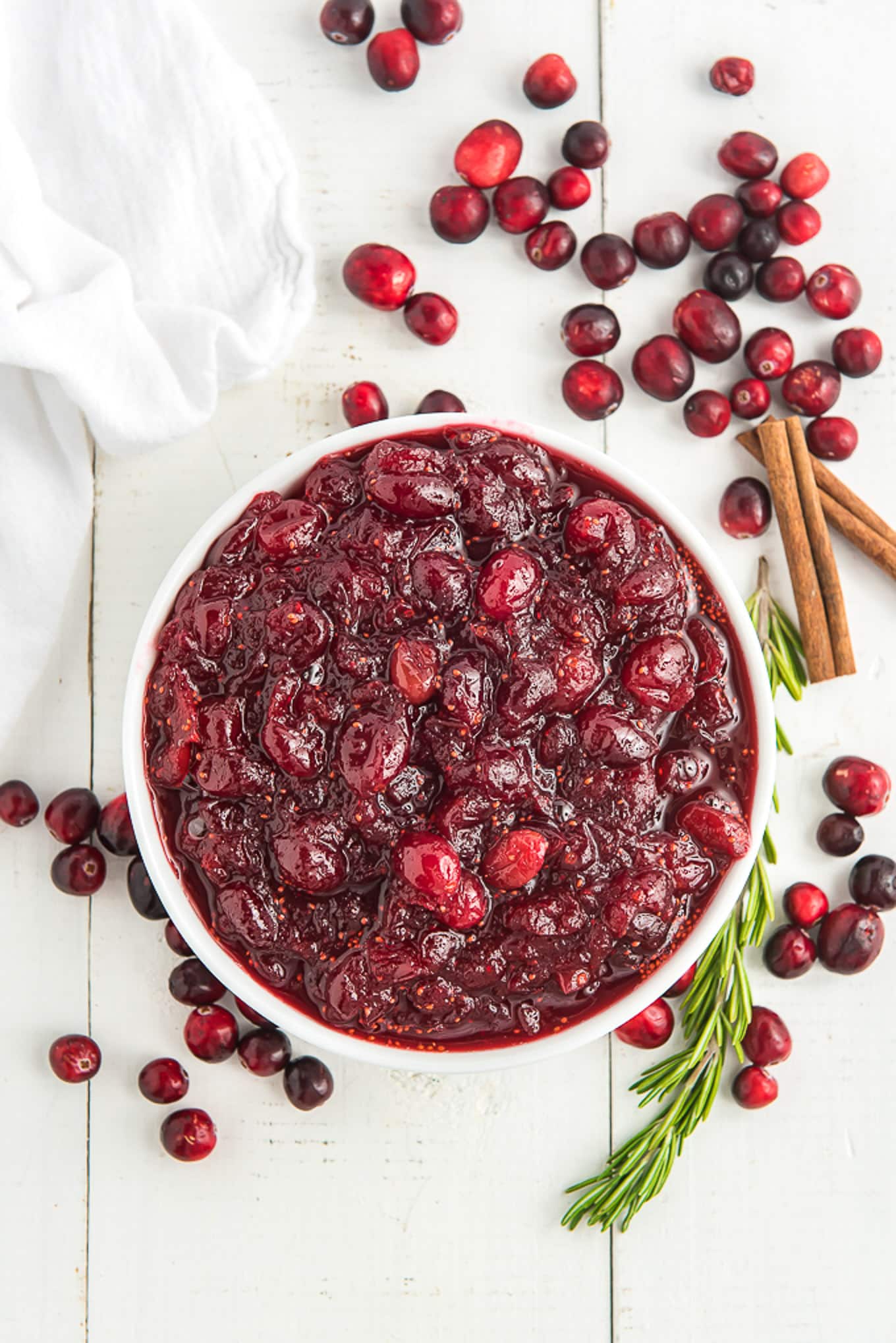 A bowl of cranberry sauce on the table with rosemary, fresh cranberries, and cinnamon sticks to the side.