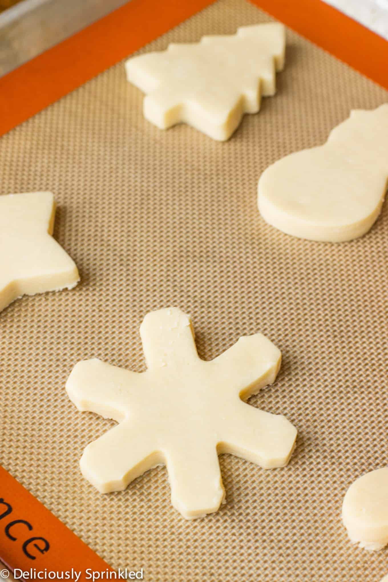 Sugar cookie cut-outs in trees, snowmen, and snowflake shapes on a silpat lined baking tray before baking.