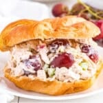 Rotisserie chicken salad on a a croissant on a plate with grapes in the back.