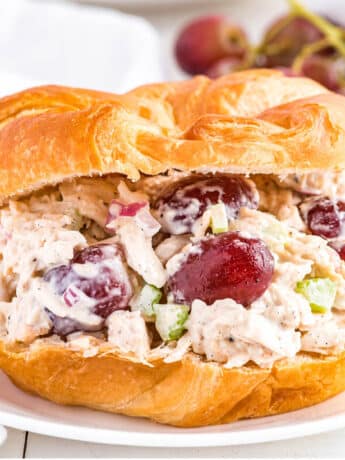 Rotisserie chicken salad on a a croissant on a plate with grapes in the back.